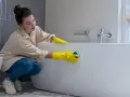 Femme en gants jaunes effectuant le nettoyage d'une baignoire acrylique avec une éponge.