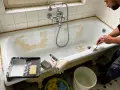 Un homme est en train de peindre une baignoire ancienne. Des outils comme un pinceau, un rouleau et de la peinture sont à proximité.