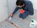 Préparation d'un ragréage sur carrelage ancien. Un ouvrier, masqué et ganté, applique un primaire d'accrochage au pinceau sur le sol carrelé.