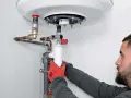 Un homme installe un ballon d'eau chaude, manipulant avec soin les tuyaux en cuivre et le siphon blanc.