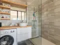 Coin buanderie dans salle de bain moderne avec lave-linge, lavabo, douche vitrée et étagères décoratives.