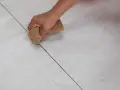 Main tenant, quelle couleur de joint pour carrelage beige ? Une main utilise une éponge pour nettoyer les joints entre des carreaux beiges clairs.