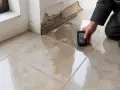 Un professionnel utilise un humidimètre pour diagnostiquer un carrelage qui se décolle à cause de l'humidité, révélant des dégâts importants.