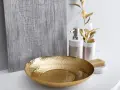 Un bol martelé doré, un distributeur de savon et une barre de savon sur un comptoir blanc, avec une plante dorée et un panneau texturé gris en arrière-plan. Une touche de luxe pour votre douche dorée.