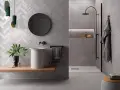 Salle de bain moderne avec murs en **grès cérame** à chevrons et texturés. Lavabo suspendu, douche à l'italienne, miroir rond et touches végétales.