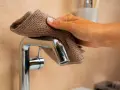 Main d'une femme nettoyant un mitigeur de douche avec un tissu marron.