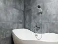 Baignoire blanche aux formes douces, adossée à un mur de carrelage gris en pose en quinconce. Douchette chromée au-dessus.