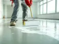 Application d'une primaire d'accrochage avant étanchéité sur un sol en béton. Un ouvrier en combinaison grise et gants rouges utilise un rouleau pour étaler le produit blanc.