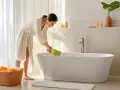 Une femme en peignoir blanc utilise une éponge verte pour nettoyer la baignoire, éliminant une tache jaune tenace.