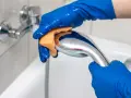Mains bleues gantées frottant un pommeau de douche chromé avec une éponge orange pour le nettoyer.