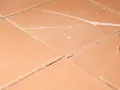 Carrelage mal posé, avec des carreaux fissurés et des joints irréguliers. Des éclats de matière blanche parsèment la surface.