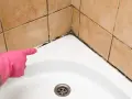 Main d'une personne portant un gant rose pointe vers le joint de douche moisi, indiquant qu'il faut refaire le joint de douche.