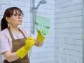 Femme avec des gants jaunes et une lingette verte en train de nettoyer le calcaire de la douche.