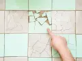 Un doigt pointe une **fissure carrelage** sur un mur de carreaux anciens, certains brisés, d'autres craquelés.