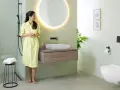 Une femme en robe jaune admire une salle de bain moderne. Le mur vert pâle est une tendance couleur mur toilette, complété par un meuble vasque en bois et un WC suspendu.