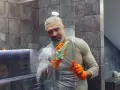 Un homme aux cheveux gris, portant des gants orange, nettoie une paroi de douche en plexiglas avec une éponge savonneuse.