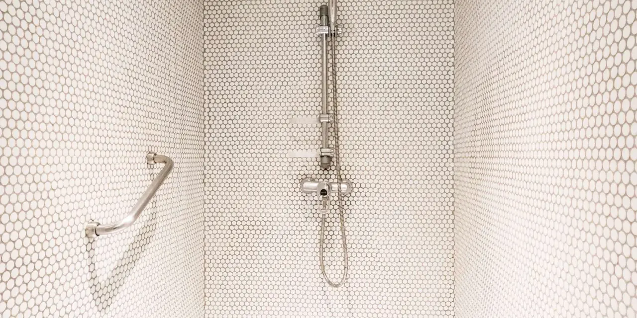 Douche moderne avec carrelage blanc en mosaïque. Une barre d'appui chromée et un pommeau de douche argenté se détachent sur le fond. Le receveur noir est discret.