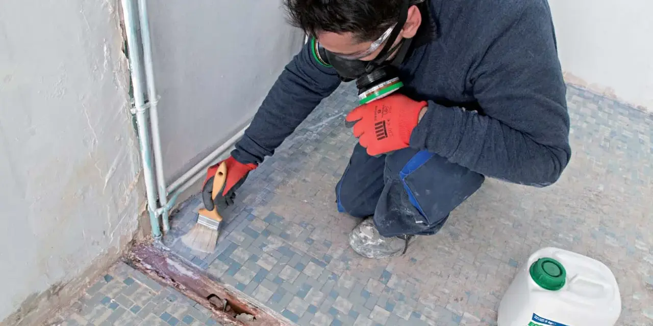 Préparation d'un ragréage sur carrelage ancien. Un ouvrier, masqué et ganté, applique un primaire d'accrochage au pinceau sur le sol carrelé.