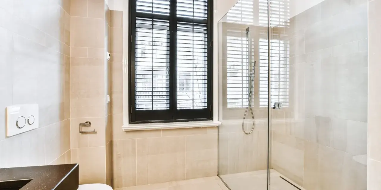 Salle de bain moderne avec douche à l'italienne, carrelage beige, fenêtre à persiennes noires et WC suspendu.