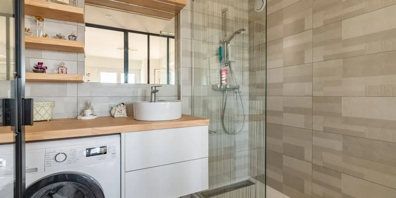 Coin buanderie dans salle de bain moderne avec lave-linge, lavabo, douche vitrée et étagères décoratives.