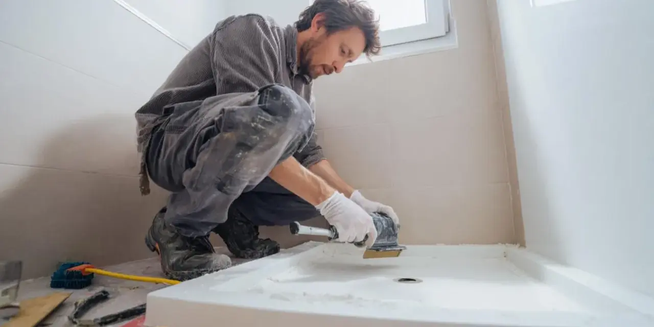 Un ouvrier prépare le sol pour installer un receveur de douche. Il utilise une ponceuse pour assurer une surface plane, essentielle pour le collage.