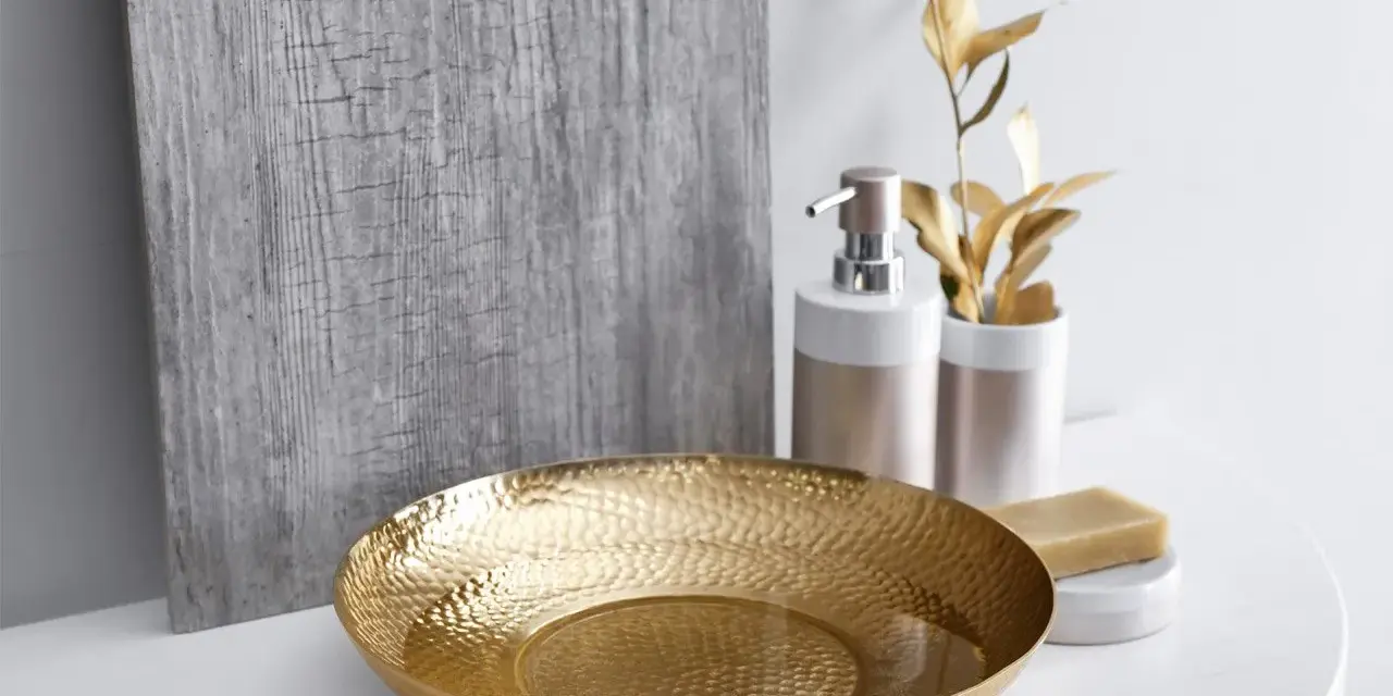 Un bol martelé doré, un distributeur de savon et une barre de savon sur un comptoir blanc, avec une plante dorée et un panneau texturé gris en arrière-plan. Une touche de luxe pour votre douche dorée.