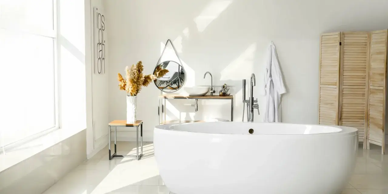 Salle de bain lumineuse avec baignoire îlot blanche, lavabo, miroir, porte-serviettes et paravent en bois. La largeur d'une baignoire est parfaite pour ce coin détente.