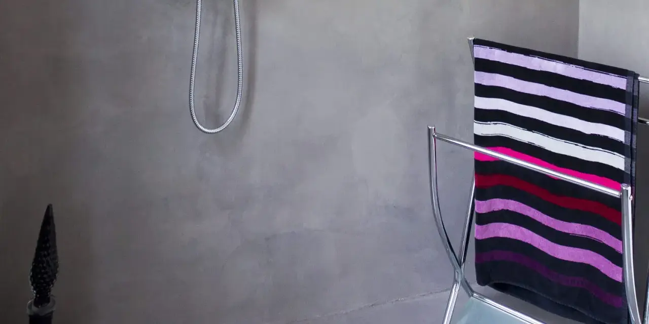 Salle de bain béton ciré aux tons gris, avec une douche chromée et un porte-serviettes orné d'une serviette rayée.