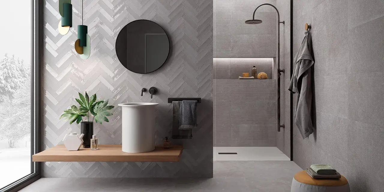 Salle de bain moderne avec murs en **grès cérame** à chevrons et texturés. Lavabo suspendu, douche à l'italienne, miroir rond et touches végétales.
