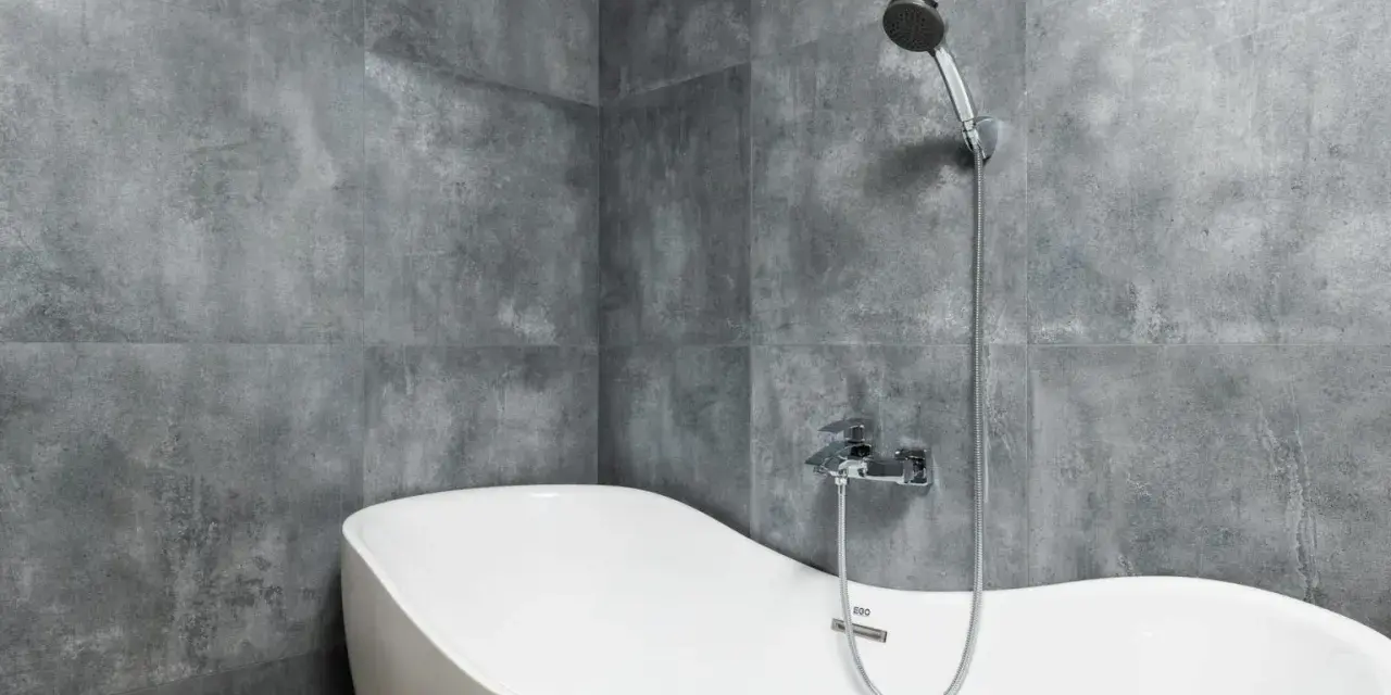 Baignoire blanche aux formes douces, adossée à un mur de carrelage gris en pose en quinconce. Douchette chromée au-dessus.