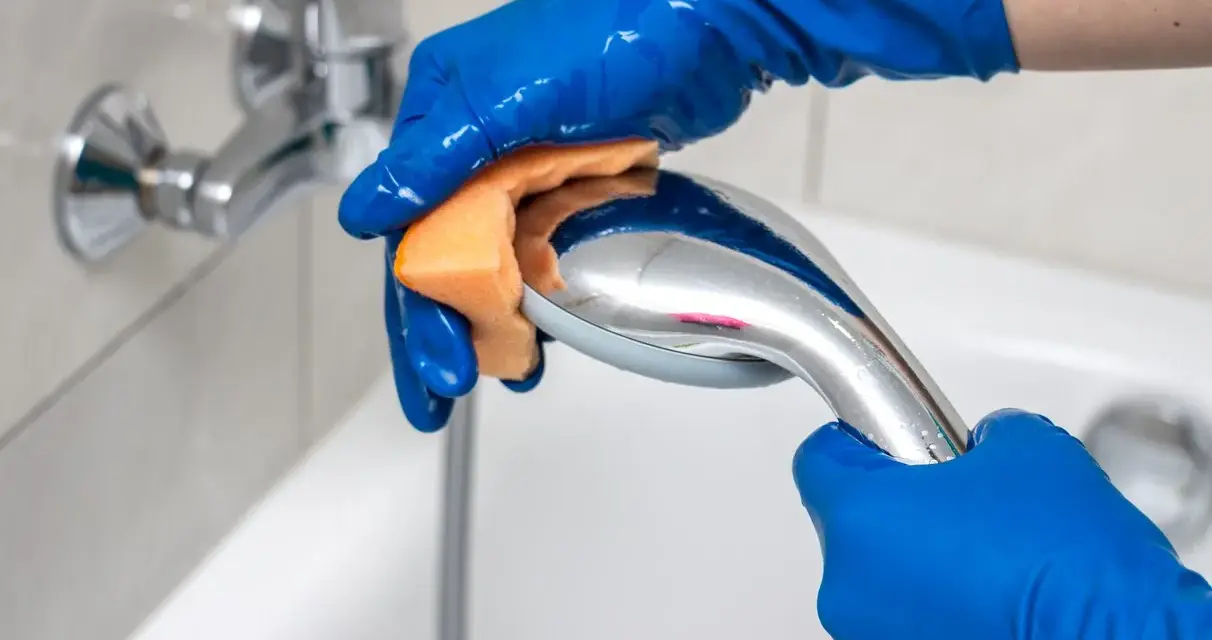 Mains bleues gantées frottant un pommeau de douche chromé avec une éponge orange pour le nettoyer.