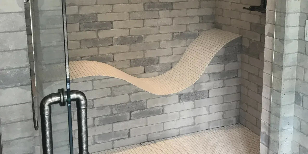 Douche moderne avec un banc ondulé intégré dans le mur de briques. La poignée de la porte en verre est en métal vieilli.