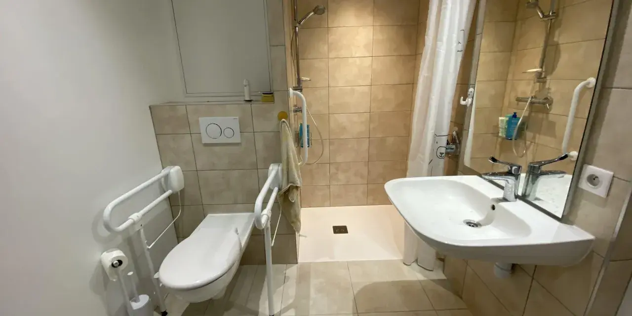 Salle de bain pmr avec douche à l'italienne, WC avec barres d'appui et lavabo. Confort et sécurité pour tous.