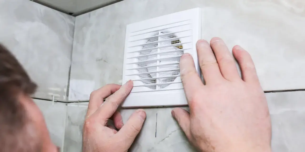 Mains d'homme ajustant une grille de VMC dans une salle de bain car une mauvaise odeur persiste.
