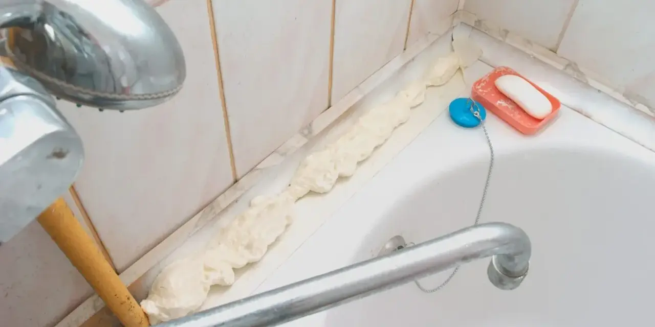 Mousse expansive appliquée pour nettoyer joint plastique porte douche. Savon et bouchon de bain à proximité.