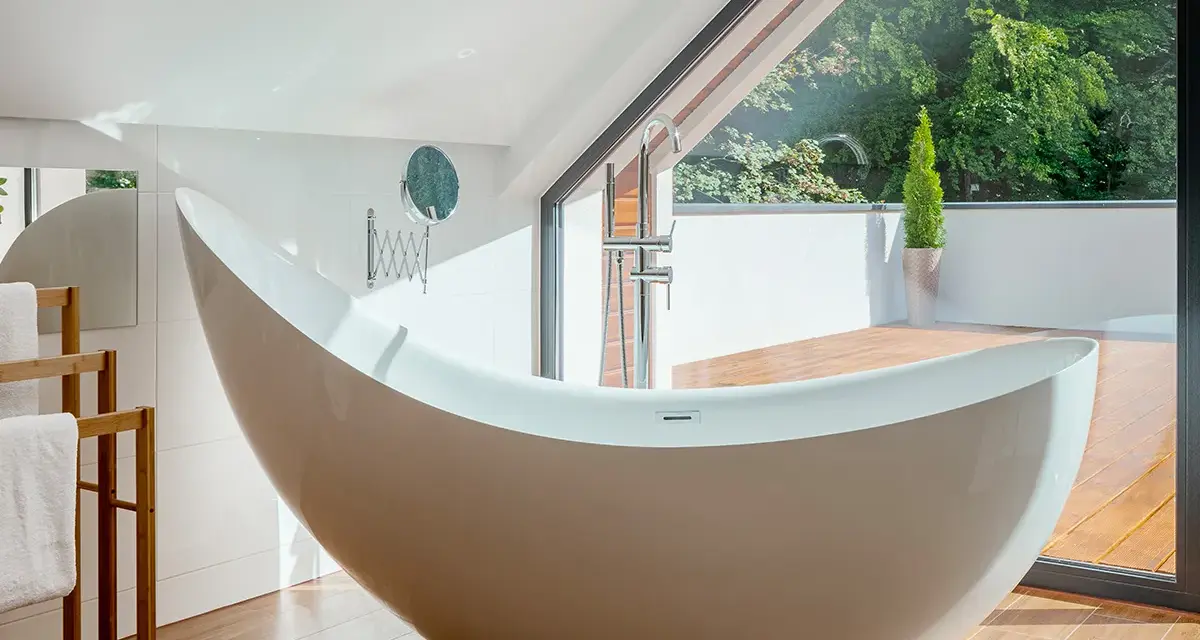 Baignoire design en matière composite, avec robinetterie chromée, devant une grande baie vitrée donnant sur un jardin luxuriant.