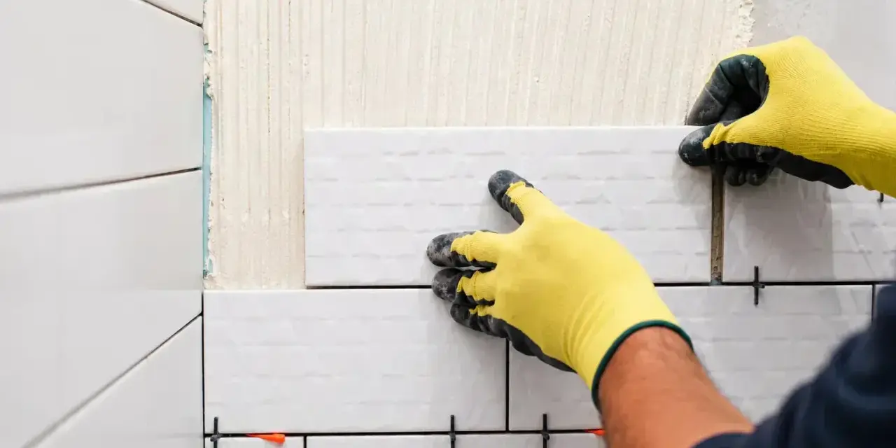 Mains gantées de jaune en train de coller du carrelage sur du carrelage, avec des croisillons pour un espacement parfait.