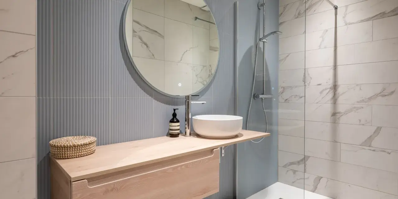 Aménagement petite salle de bain : meuble suspendu en bois clair, vasque ronde, miroir rond, douche à l'italienne.
