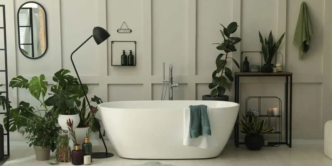 Une salle de bain baignée de lumière, avec une baignoire blanche, de nombreuses plantes vertes qui prospèrent dans l'humidité, et un miroir ovale.
