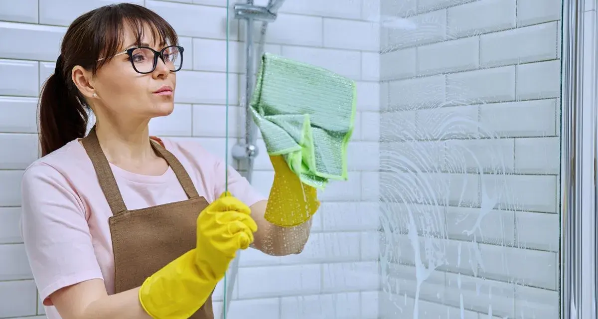 Femme avec des gants jaunes et une lingette verte en train de nettoyer le calcaire de la douche.