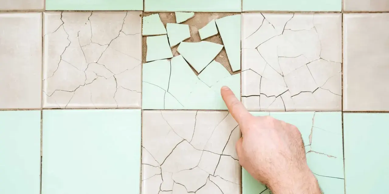 Un doigt pointe une **fissure carrelage** sur un mur de carreaux anciens, certains brisés, d'autres craquelés.