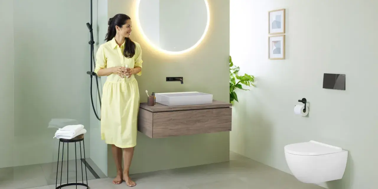 Une femme en robe jaune admire une salle de bain moderne. Le mur vert pâle est une tendance couleur mur toilette, complété par un meuble vasque en bois et un WC suspendu.