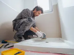 Un ouvrier prépare le sol pour installer un receveur de douche. Il utilise une ponceuse pour assurer une surface plane, essentielle pour le collage.