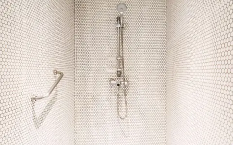 Douche moderne avec carrelage blanc en mosaïque. Une barre d'appui chromée et un pommeau de douche argenté se détachent sur le fond. Le receveur noir est discret.