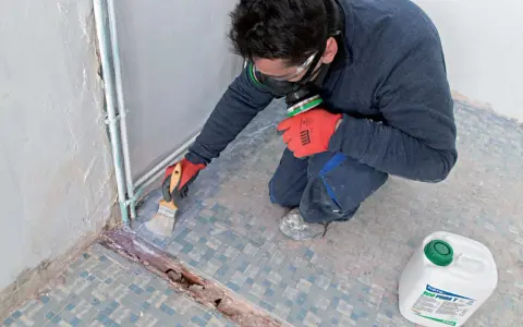 Préparation d'un ragréage sur carrelage ancien. Un ouvrier, masqué et ganté, applique un primaire d'accrochage au pinceau sur le sol carrelé.