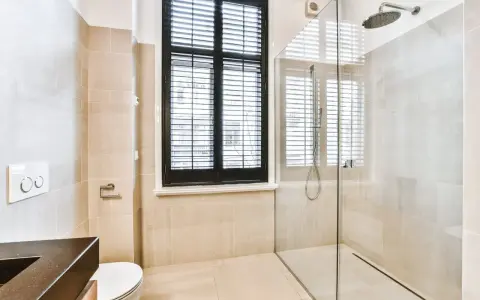 Salle de bain moderne avec douche à l'italienne, carrelage beige, fenêtre à persiennes noires et WC suspendu.