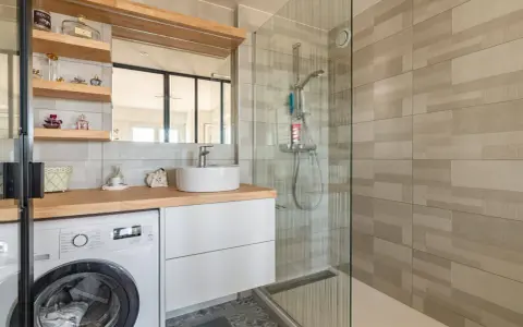 Coin buanderie dans salle de bain moderne avec lave-linge, lavabo, douche vitrée et étagères décoratives.