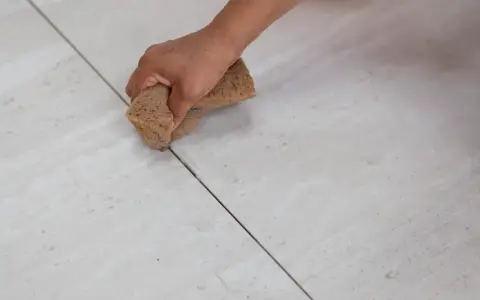 Main tenant, quelle couleur de joint pour carrelage beige ? Une main utilise une éponge pour nettoyer les joints entre des carreaux beiges clairs.