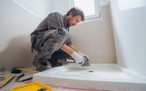 Un ouvrier prépare le sol pour installer un receveur de douche. Il utilise une ponceuse pour assurer une surface plane, essentielle pour le collage.