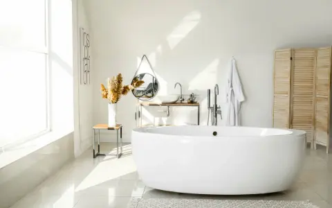 Salle de bain lumineuse avec baignoire îlot blanche, lavabo, miroir, porte-serviettes et paravent en bois. La largeur d'une baignoire est parfaite pour ce coin détente.